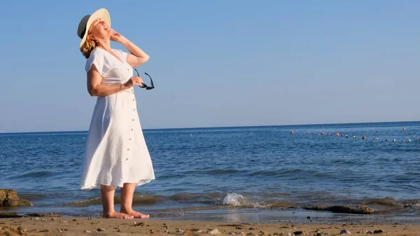 Женщина в соломенной шляпе и белом платье снимает солнечные очки и наслаждается солнцем, прогуливаясь по берегу моря и любуясь голубым морем в солнечный летний день, наслаждаясь свободой и отдыхом. При этом — стоковое фото