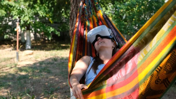 Mulher engraçada vestindo óculos de realidade virtual repousa em uma rede perto de uma árvore, imaginando um sonho tornado realidade, um mundo inventado. O conceito de obter experiência usando óculos de fone de ouvido VR de virtual — Fotografia de Stock
