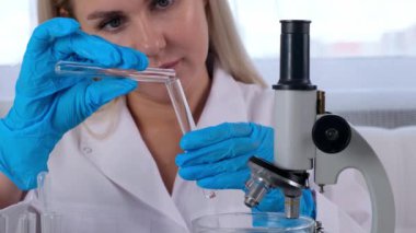 Tıbbi giysi ve koruyucu eldiven giymiş kadın bilim adamı laboratuvar asistanı laboratuarda mikroskop altında çalışmak üzere test tüpleriyle numuneler üzerinde araştırma yapıyor...
