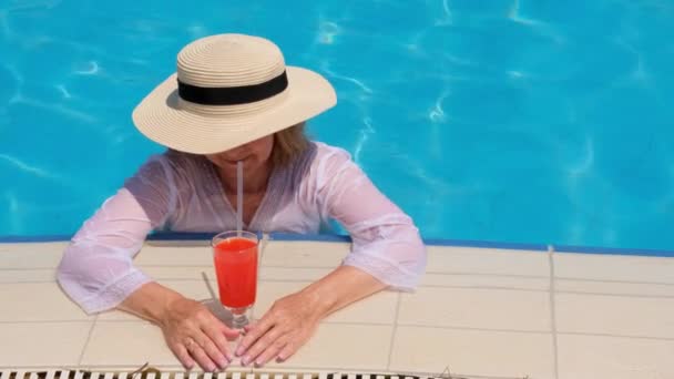 Lächelnde blonde Frau mit Strohhut entspannt sich und trinkt an einem sonnigen Sommertag in der Nähe des Swimmingpools einen orangefarbenen Cocktail. Sommerliche Stimmung, Urlaub, all inclusive — Stockvideo