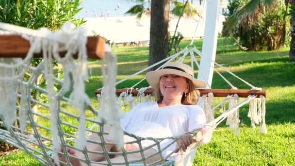 Starsza kobieta w słomkowym kapeluszu uśmiechnięta szczęśliwa relaksująca się na hamaku ciesząca się świeżym powietrzem na tarasie wokół palm w pobliżu morza. Koncepcja stylu życia seniora — Wideo stockowe