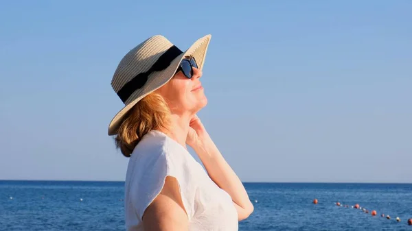 Счастливая зрелая женщина 50 лет, одетая в белое платье, соломенный пляж и солнечные очки на пляже у берега моря. Концепция отдыха, релаксации, пенсионного возраста — стоковое фото