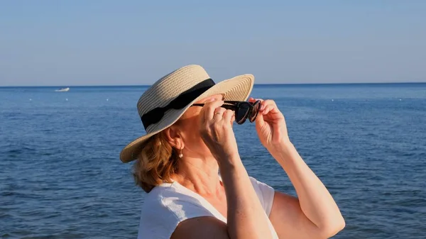 Портрет зрелой женщины, 50 лет, в соломенной шляпе и солнечных очках на синем морском фоне. Лето, отпуск, отпуск, активные пенсионеры — стоковое фото