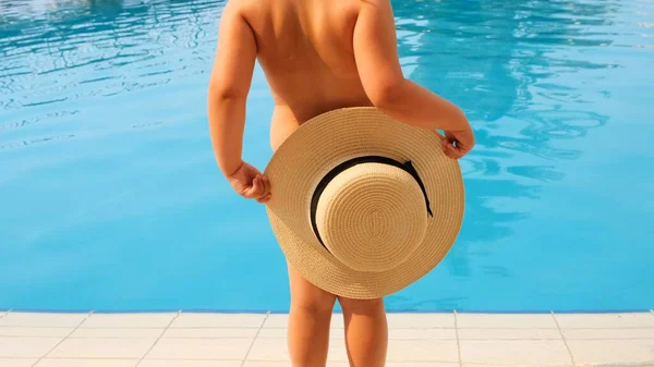 Мила дівчинка 4 роки з солом'яним капелюхом танцює і дивиться на блакитне море або басейн, вид ззаду, впритул. Дитинство, стосунки, концепція літніх канікул. вибірковий фокус на капелюсі . — стокове фото