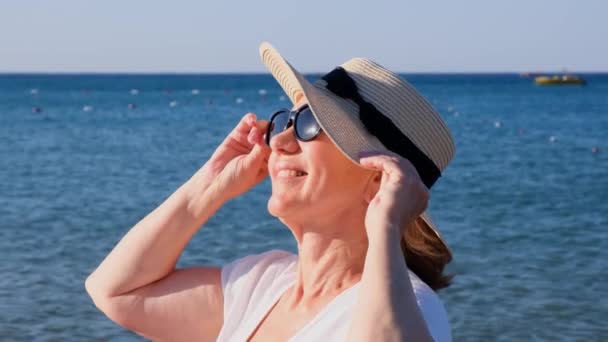 Glückliche reife Frau 50 Jahre alt in weißem Kleid, Strohstrand und Sonnenbrille am Strand in der Nähe der Küste. Urlaubskonzept, Entspannung, Rentenalter — Stockvideo