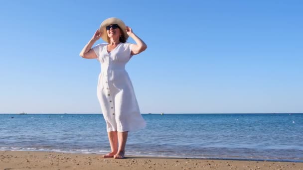 Dojrzała kobieta w okularach przeciwsłonecznych w słomkowym kapeluszu i białej sukience spaceruje wzdłuż błękitnego wybrzeża morza w słoneczny letni dzień, ciesząc się wolnością i relaksem. Koncepcja życia codziennego seniora — Wideo stockowe