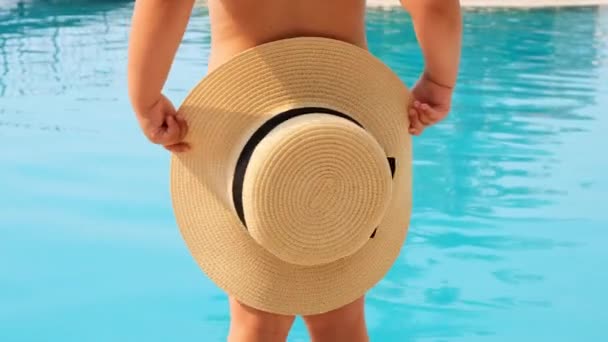 Sweet little girl 4 years old with a straw hat dances and looks at the blue sea or pool, back view, close-up. Childhood, relationship, summer vacation concept. selective focus on the hat. — Stock Video