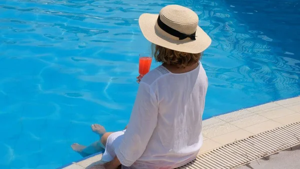 プールの近くでリラックスした藁帽子の女性ピンクのカクテルを飲んで水に足をぶら下げて。夏気分のコンセプト — ストック写真