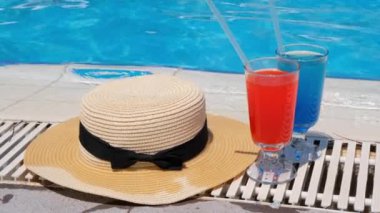 İki renkli kokteyl bardağı pembe ve mavi, yüzme havuzunun kenarında güneş şapkası, yakın çekim. Tatil konsepti, otel rahatlığı, her şey dahil.