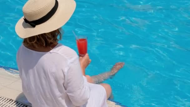 Frau mit Strohhut entspannt sich in der Nähe des Swimmingpools, trinkt einen rosa Cocktail und lässt ihre Beine im Wasser baumeln. Sommerliche Stimmung — Stockvideo