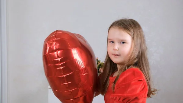Fetiță drăguță într-o rochie roșie care ține baloane roșii în formă de inimă în mâinile ei conceptul de zi Valentine — Fotografie, imagine de stoc