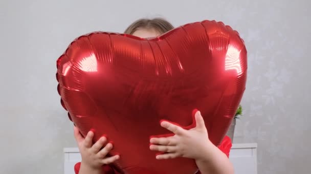 Niña linda en un vestido rojo se esconde detrás de un globo en forma de corazón rojo. concepto de día de San Valentín — Vídeos de Stock