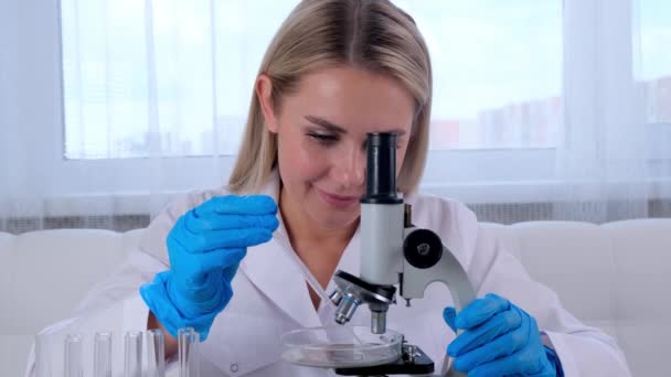 Vrouwelijke onderzoeker scheikundige wetenschapper in een medisch pak en beschermende handschoenen doet onderzoek naar monsters met een micropipette en reageerbuizen voor het werken onder een microscoop in een laboratorium. — Stockvideo