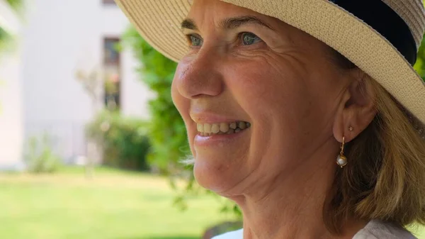 Portrét dospělé ženy ve věku 55-60 let se zubatým úsměvem na slunci chránícím slamáku, užívající si svůj čas na pozadí zelené zahrady. close-up. — Stock fotografie