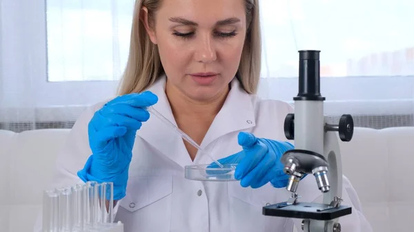 Uma mulher cientista química de pesquisa em um terno médico e luvas de proteção realiza pesquisas em amostras com uma micropipeta e tubos de ensaio para trabalhar sob um microscópio em um laboratório.. — Fotografia de Stock
