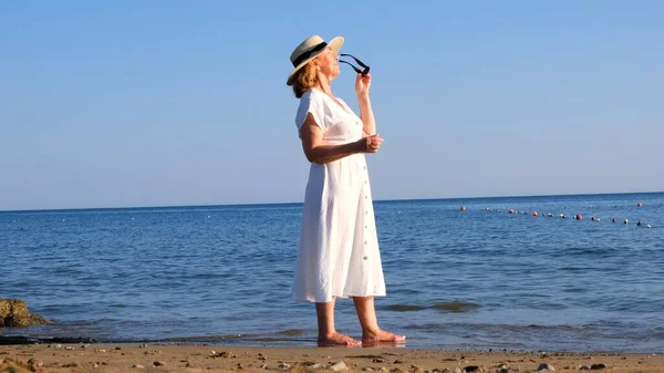 Женщина в соломенной шляпе и белом платье гуляет по берегу моря в солнечных очках и любовается голубым морем в солнечный летний день, наслаждаясь свободой и отдыхом. Концепция пожилого человека — стоковое фото
