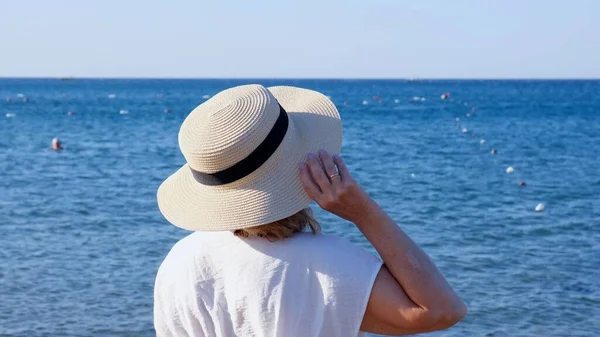 Вид сзади на женщину в соломенной шляпе и белом платье, смотрящую на синее море, крупным планом. Концепция отдыха, солнцезащитного крема — стоковое фото