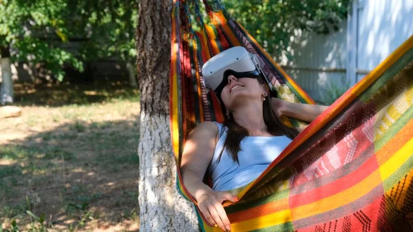 Mulher alegre vestindo óculos de realidade virtual repousa em uma rede perto de uma árvore, imaginando um sonho tornado realidade, um mundo inventado. — Fotografia de Stock