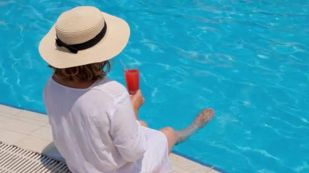Mujer en un sombrero de paja se relaja cerca de la piscina, bebe un cóctel rosa y cuelga sus piernas en el agua. Concepto de humor de verano — Vídeos de Stock