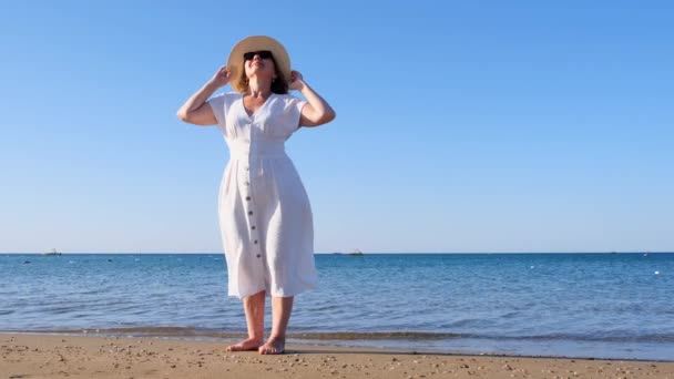 Dojrzała kobieta w słomkowym kapeluszu i białej sukience ciesząca się słońcem spacerując wzdłuż błękitnego morza w słoneczny letni dzień, ciesząc się wolnością i relaksem. Koncepcja życia codziennego seniora — Wideo stockowe
