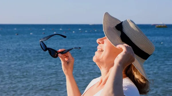 Счастливая 50-летняя женщина в соломенной шляпе и солнцезащитных очках, наслаждающаяся солнцем на фоне синего моря. Лето, отпуск, отпуск, активные пенсионеры — стоковое фото
