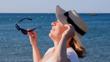 Hasır şapkalı ve güneş gözlüklü mutlu 50 yaşındaki kadın mavi deniz arka planında güneşin tadını çıkarıyor. Yaz, tatil, tatil, aktif emekliler