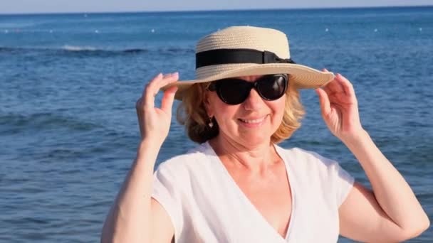 Piękna 50-letnia kobieta w słomkowym kapeluszu i okularach przeciwsłonecznych na błękitnym tle morza. Lato, wakacje, wakacje, aktywni emeryci — Wideo stockowe