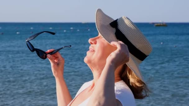 Wanita berusia 50 tahun yang bahagia dengan topi jerami dan kacamata hitam menikmati matahari dengan latar belakang laut biru. Musim panas, liburan, liburan, pensiunan aktif — Stok Video