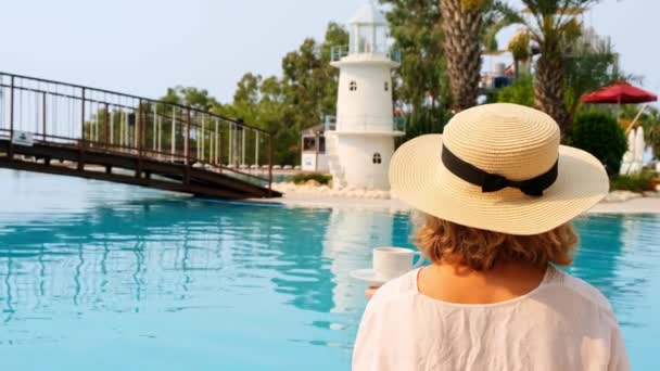 Kobieta w słomkowym kapeluszu w białej sukience relaksująca się przy basenie z filiżanką kawy, koncepcja wakacji, dzień dobry. kobieta siedząca przy basenie — Wideo stockowe