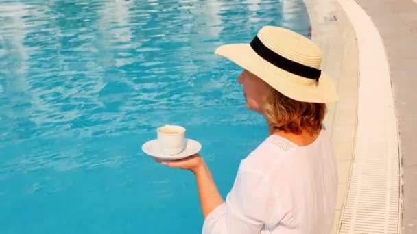 Жінка 50 років поснідала біля басейну в солом'яному капелюсі в білій сукні. жінка сидить біля басейну з чашкою кави. доброго ранку і денного планування — стокове відео