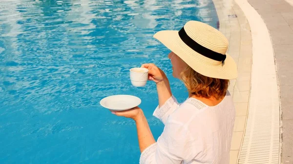 Ідеальна концепція розкоші. ранкова жінка в солом'яному капелюсі п'є каву біля блакитного басейну — стокове фото