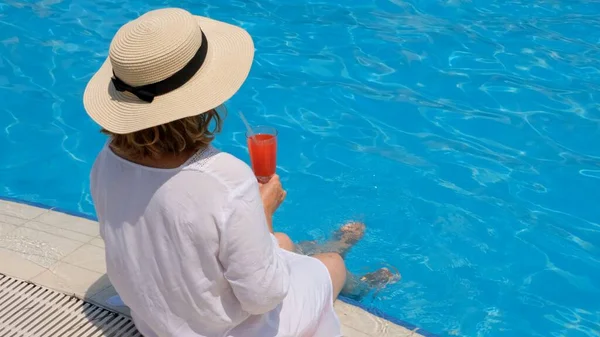 プールの近くでリラックスした藁帽子の女性ピンクのカクテルを飲んで水に足をぶら下げて。夏気分のコンセプト — ストック写真