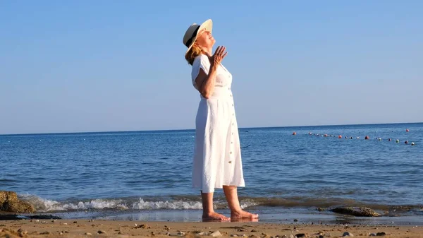 Зрелая женщина в соломенной шляпе и белом платье наслаждается солнцем, гуляя вдоль синего побережья моря в солнечный летний день, наслаждаясь свободой и отдыхом. Концепция повседневной жизни пожилых людей — стоковое фото