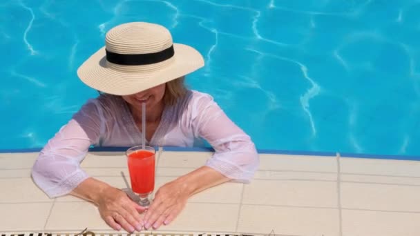 Femme blonde souriante en chapeau de paille se détend et boit cocktail orange près de la piscine par une journée d'été ensoleillée. Concept d'ambiance estivale, vacances, tout compris — Video