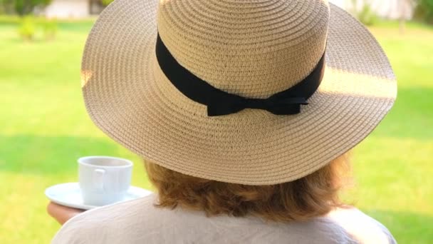 Vista posteriore di una donna con un cappello di paglia con una tazza bianca di bevanda calda di caffè nel suo giardino sulla terrazza in estate con una giornata di sole. Concetto mattutino, divertimento mattutino, sintonia per un giorno felice — Video Stock