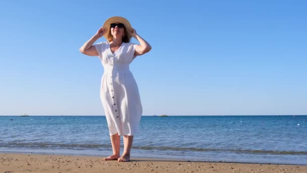Mature woman in sunglasses in a straw hat and white dress walks along the blue sea coast on a sunny summer day, enjoying freedom and relaxation. The concept of a senior citizen everyday life — Stock Video