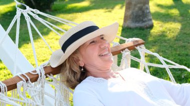 Hasır şapkalı yaşlı kadın mutlu bir şekilde hamakta rahatlıyor ve deniz kenarındaki palmiye ağaçlarının etrafındaki terasta temiz havanın tadını çıkarıyor. Yaşlılar yaşam tarzı konsepti