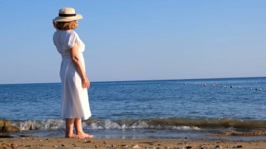 Güneşli bir yaz gününde mavi deniz kıyısında yürürken hasır şapkalı ve beyaz elbiseli olgun bir kadının arkasına bakmak özgürlük ve rahatlığın tadını çıkarıyor. Emeklilerin tipik hayatı kavramı.