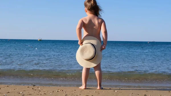 Забавная маленькая девочка 3 года в соломенной шляпе смотрит на море, вид сзади, крупный план. Детство, отношения, концепция летних каникул. Избирательный акцент на шляпе — стоковое фото