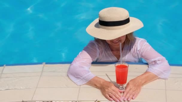 Mujer rubia sonriente en sombrero de paja relaja y bebe cóctel naranja cerca de la piscina en un día soleado de verano. Concepto de humor de verano, vacaciones, todo incluido — Vídeo de stock