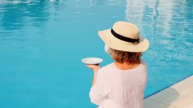 Hasır şapkalı bir kadın havuz kenarında dinlenen beyaz bir elbise giyiyor bir fincan kahve, tatil konsepti, günaydın. Havuz kenarında oturan kadın.