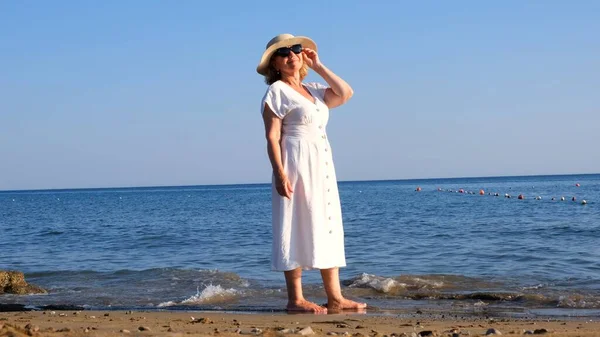 Donna matura in occhiali da sole con cappello di paglia e abito bianco cammina lungo la costa blu in una giornata estiva soleggiata, godendo di libertà e relax. Il concetto di vita quotidiana degli anziani — Foto Stock