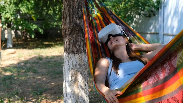 Mulher alegre vestindo óculos de realidade virtual repousa em uma rede perto de uma árvore, imaginando um sonho tornado realidade, um mundo inventado. — Fotografia de Stock
