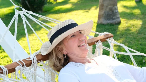 Hasır şapkalı yaşlı kadın palmiye ağaçlarının etrafındaki bir hamakta dinleniyor ve huzur ve rahatlığın tadını çıkarıyor. Yaşlılar yaşam tarzı konsepti