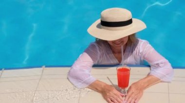Hasır şapkalı sarışın kadın güneşli bir yaz gününde yüzme havuzunda turuncu kokteyl içiyor. Tatil konsepti, hepsi dahil.