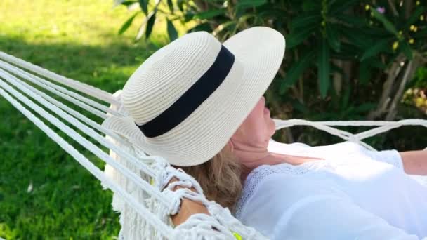 Fêmea em um chapéu de palha está descansando em uma rede ao redor das palmeiras e desfrutando da tranquilidade e relaxamento. Conceito de férias — Vídeo de Stock