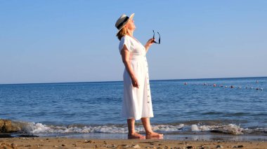 Hasır şapkalı ve beyaz elbiseli bir kadın deniz kıyısında yürür güneş gözlüğü takar ve güneşli bir yaz gününde mavi denize hayran kalır, özgürlüğün ve rahatlığın tadını çıkarır. Yaşlı vatandaş konsepti