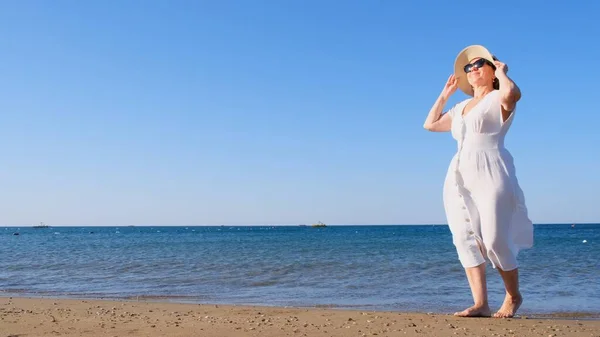Зрелая женщина в солнечных очках в соломенной шляпе и белом платье гуляет по синему морскому побережью в солнечный летний день, наслаждаясь свободой и отдыхом. Концепция повседневной жизни пожилых людей — стоковое фото