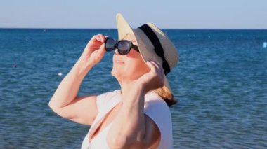Olgun bir kadının portresi, 50 yaşında, hasır şapka ve güneş gözlüğü takıyor mavi bir deniz arka planında. Yaz, tatil, tatil, aktif emekliler