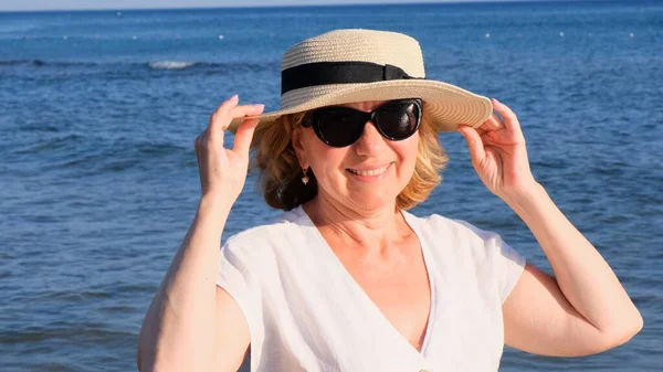 Krásná 50 letá žena v slamáku a sluneční brýle na modrém mořském pozadí. Léto, dovolená, dovolená, aktivní důchodci — Stock fotografie